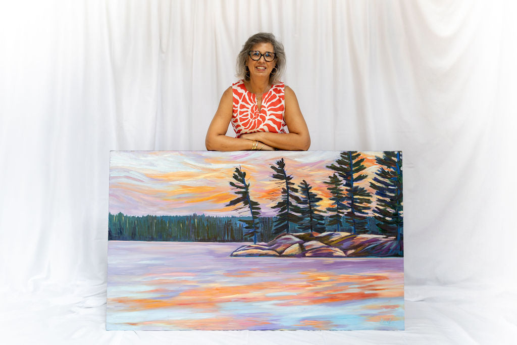 Woman standing behind a painting of a sunset over water with trees.