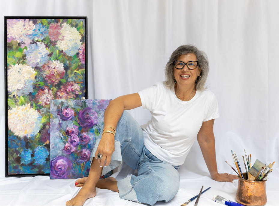 Woman sitting on a bed with floral paintings and art supplies around her